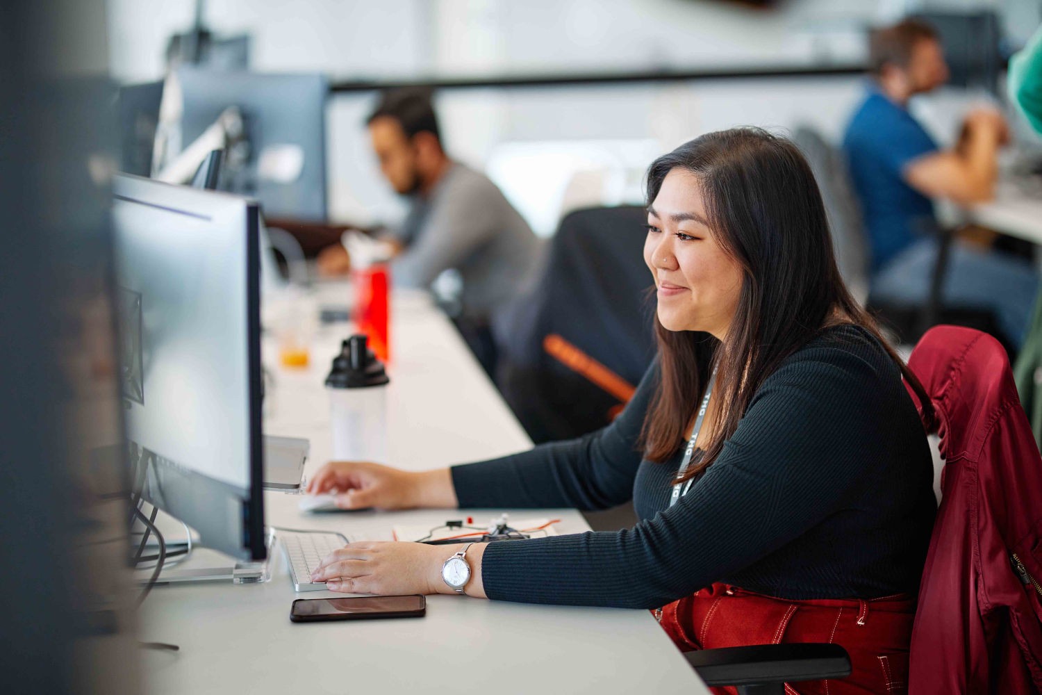 Brunette female wearing a black top and red trousers sat at her desk typing and smiling