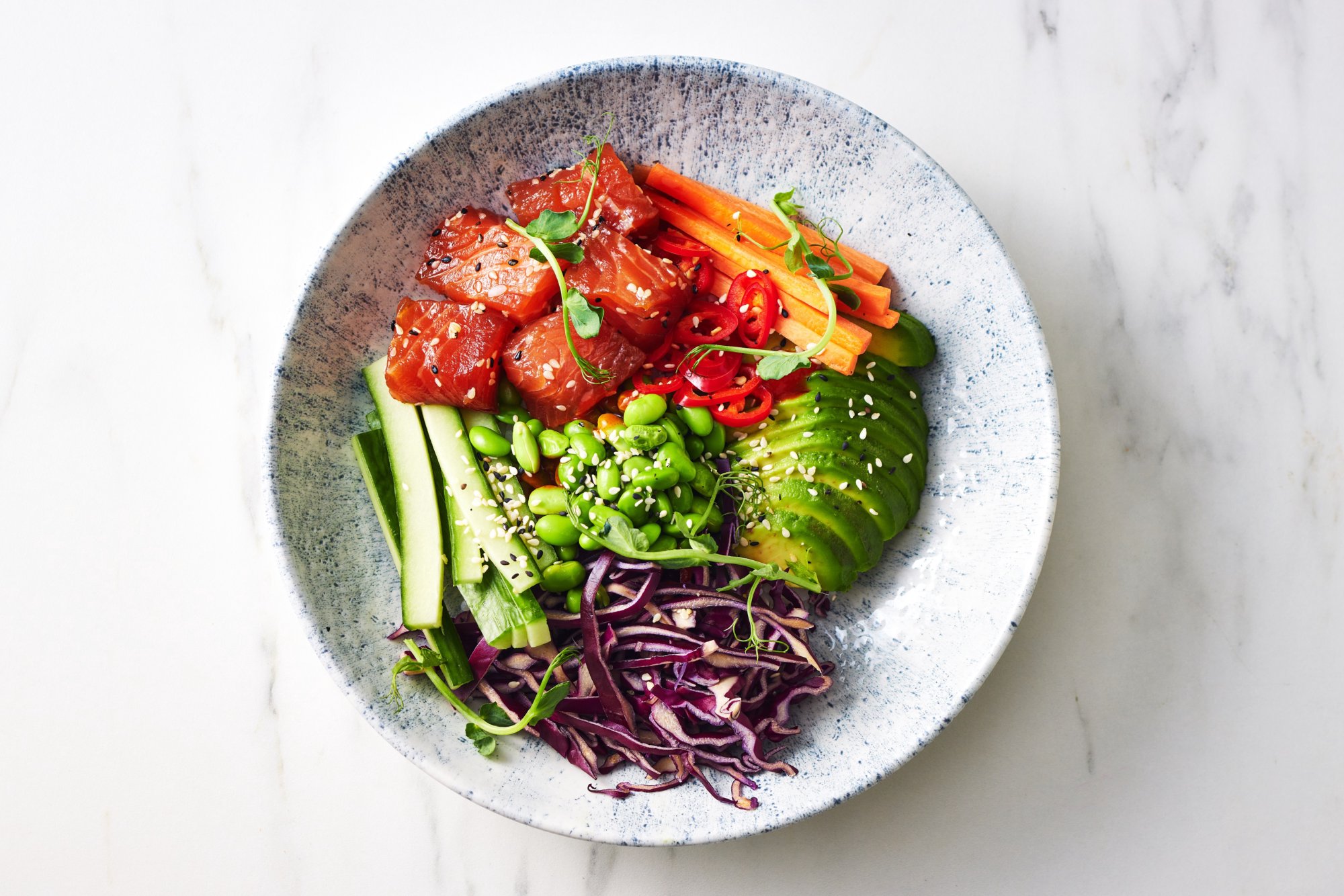 Salmon with green vegetables, carrot, cucumber and avocado in a blue textured bowl on a white marble counter top