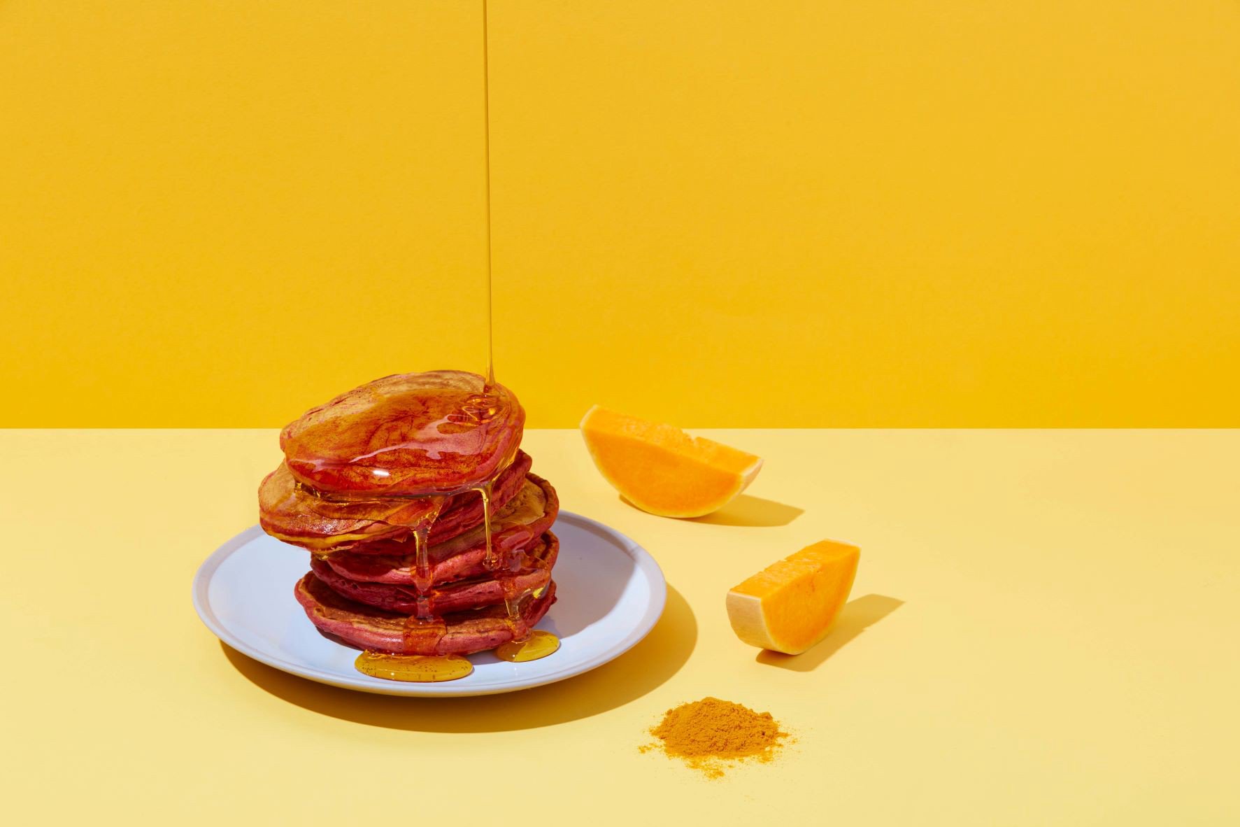 Stack of pancakes with syrup on yellow plate surrounded by two squash wedges on yellow background